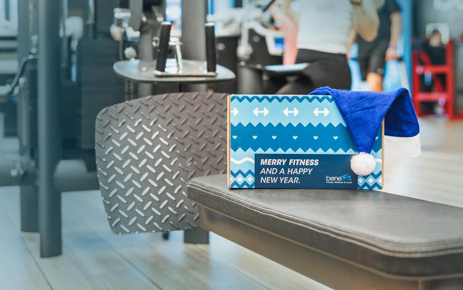 Die Merry Fitnessbox auf einer Langhantelbank im Fitnessstudio. Im Hintergrund sieht man Menschen, die Fitnesstraining machen. Auf der Box liegt eine blaue Weihnachtsmütze.