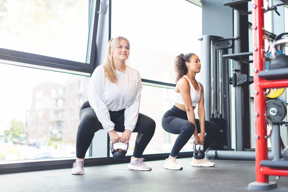 Zwei Frauen trainieren im Fitnessstudio und führen Kniebeugen mit Kettlebells vor großen Fenstern aus.