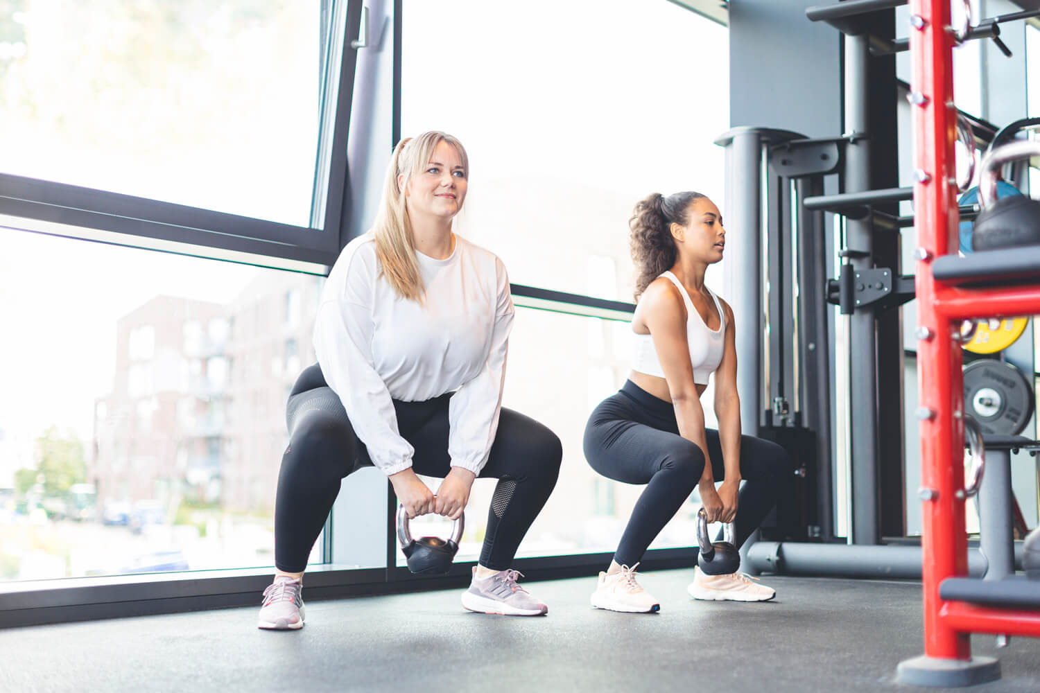 Zwei Frauen trainieren im Fitnessstudio und führen Kniebeugen mit Kettlebells vor großen Fenstern aus.