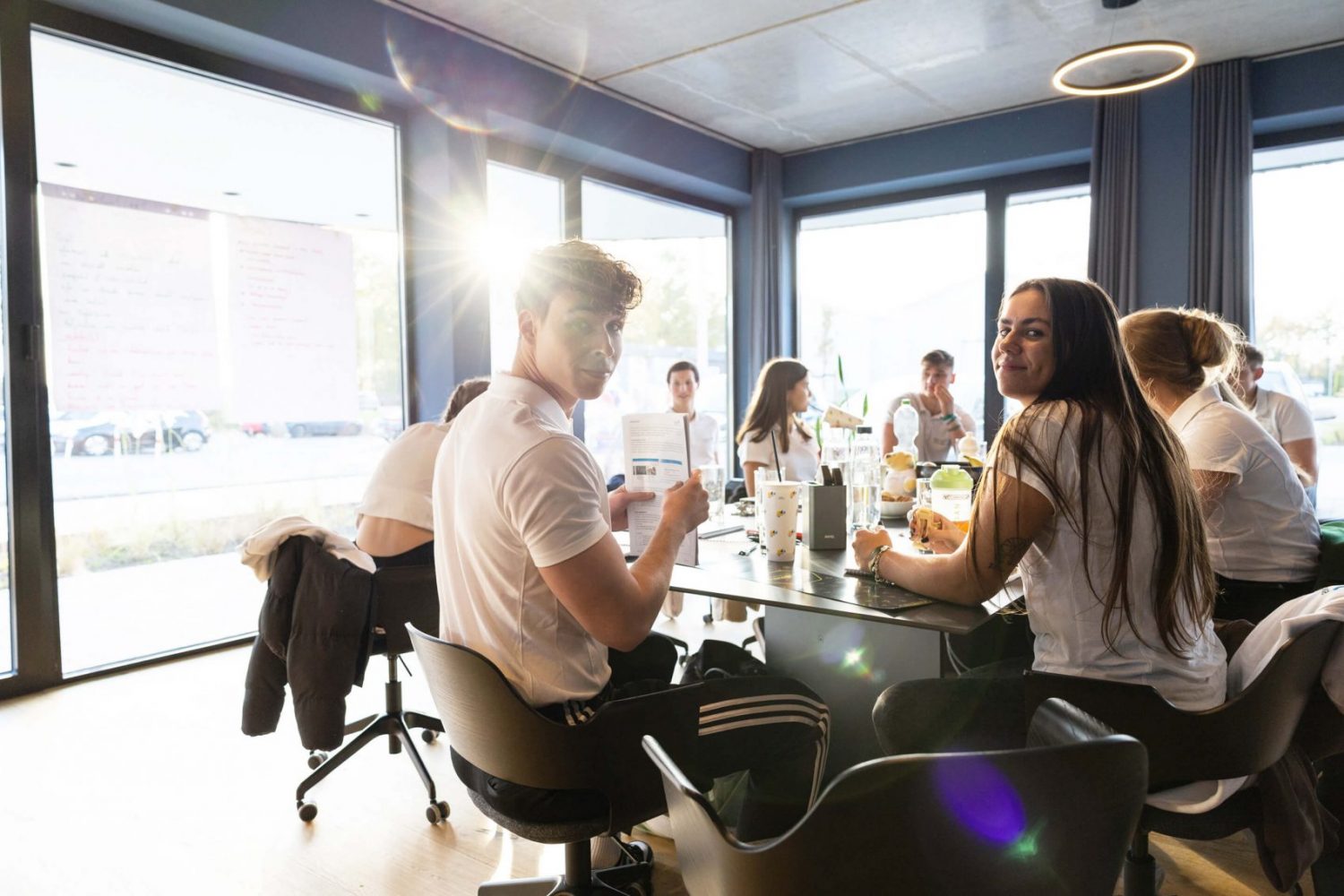 Gruppe junger Erwachsener sitzt gemeinsam an einem Tisch in einem hellen Raum mit großen Fenstern und arbeitet oder lernt zusammen; zwei Personen schauen lächelnd in die Kamera.