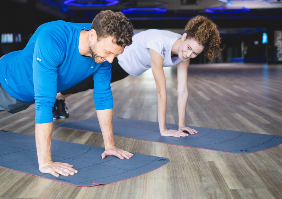 Mann und Frau machen gemeinsam Liegestütze nebeneinander auf Fitness‑Matten in einem hellen Trainingsraum, sie lächeln und wirken motiviert beim Workout.