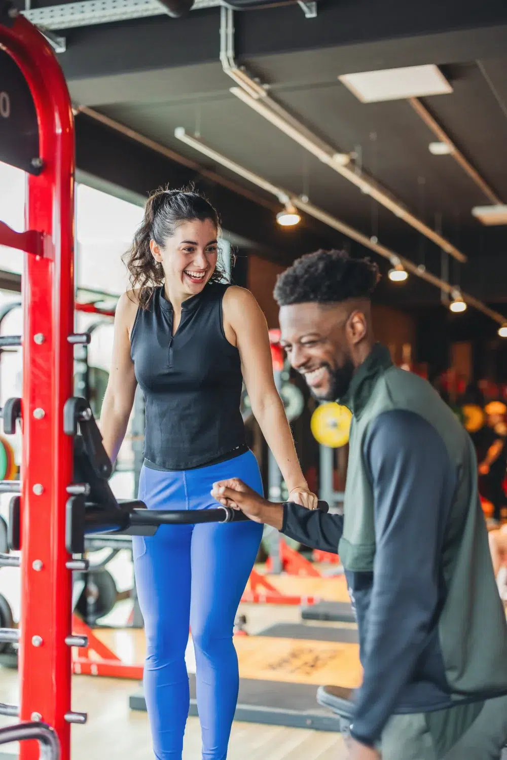 Junge Frau und junger Mann trainieren gemeinsam an einem Fitnessgerät im Fitnessstudio, beide lachen und haben Spaß.