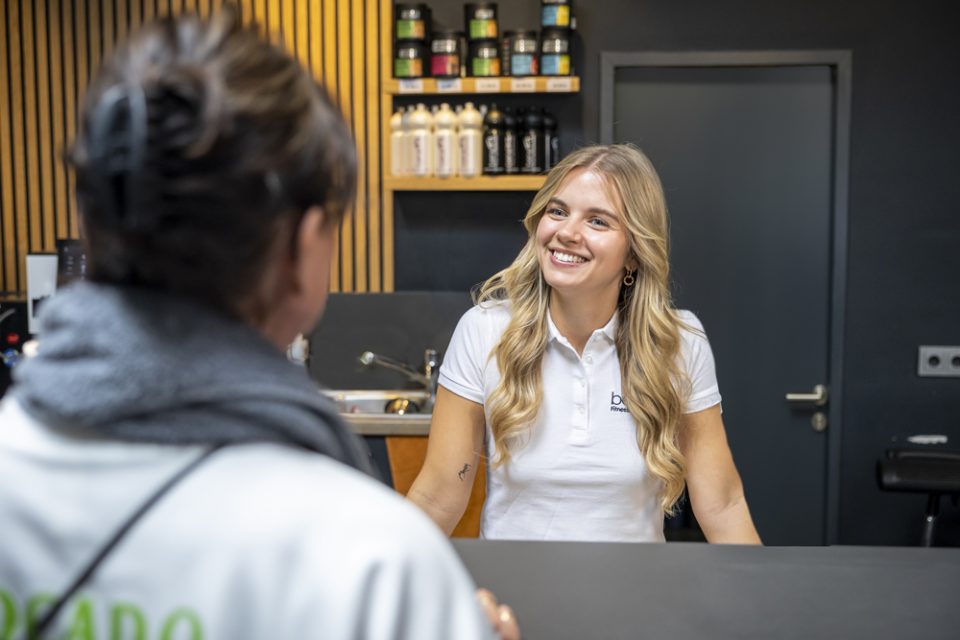 Eine beneFit-Mitarbeiterin in weißem Poloshirt mit beneFit-Logo steht hinter einem Willkommenstresen im beneFit Fitnessstudio. Im Vordergrund steht ein beneFit-Mitglied. Die beiden lächeln sich an.