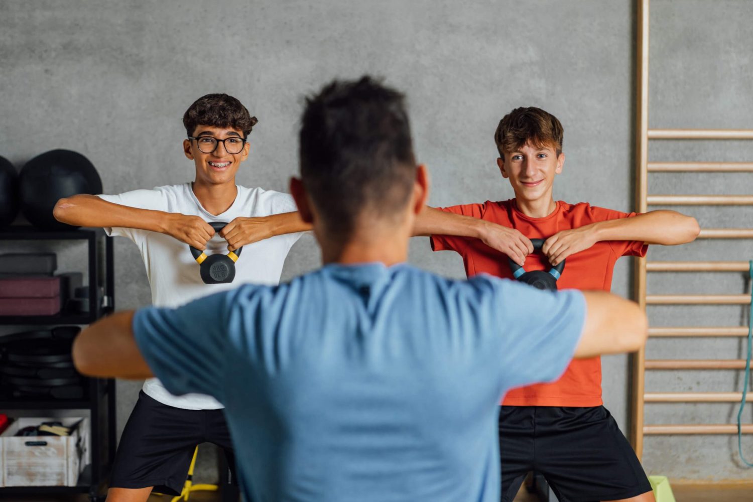 Zwei Jugendliche und ein erwachsener Trainer trainieren gemeinsam an Kettlebells im Fitnessstudio.